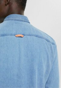 Camisa vaquera de color azul claro, con un cuello estándar y una pestaña de acento naranja en la parte trasera, con costuras visibles y tela texturizada.