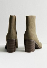 Bottes chevilles en suède vert avec une fermeture éclair sur le côté, bout rond et un talon en bloc marron côtelé. Texture lisse et design minimaliste.