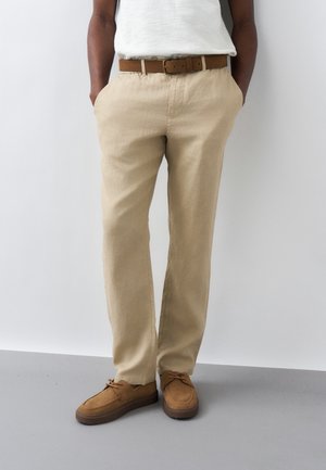 Άτομο που φοράει μπεζ παντελόνι κανονικής γραμμής, καφέ ζώνη και ταμπά σουέτ παπούτσια με κορδόνια, στέκεται με τα χέρια στις τσέπες μπροστά σε ένα λευκό τοίχο.