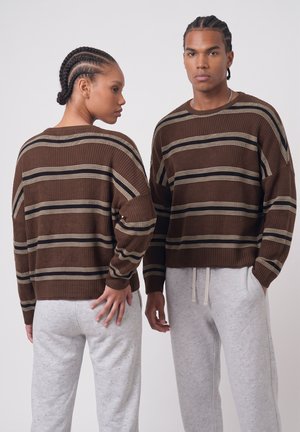 Deux personnes aux cheveux tressés portent des pulls marron rayés assortis et des pantalons de survêtement gris clair, l'une de face et l'autre de dos.