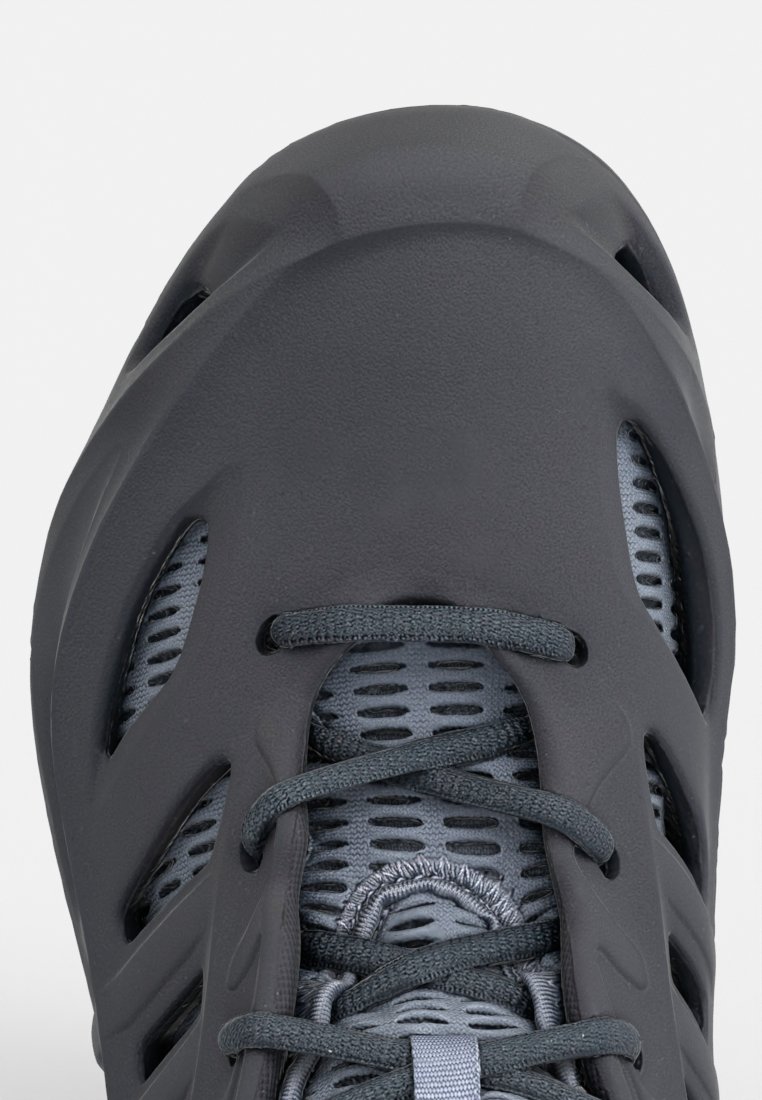 Zapatilla atlética gris con una parte superior texturizada, paneles de malla para transpirabilidad y cordones en contraste. Presenta una punta redondeada y un diseño aerodinámico.