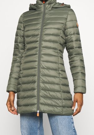 Manteau matelassé vert olive avec capuche, fermeture éclair devant et tirettes orange, porté par-dessus un jean bleu clair.
