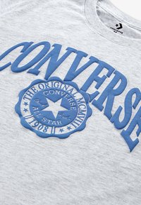 Camiseta de algodón gris con el atrevido texto en azul "CONVERSE" y un logo redondo con una estrella, que detalla "The Original All Star", establecida en 1908.