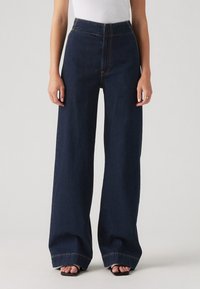 Mujer que lleva unos pantalones anchos de cintura alta en azul oscuro, combinados con sandalias negras de punta cuadrada y una blusa blanca, de pie contra un fondo liso.