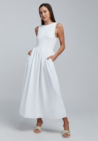 Balta midi suknelė su raukšlėtu liemeniu, be rankovių, su šoninėmis kišenėmis. Lygus audinys, lengvai krentanti suknelės apačia ir paprasta silueto forma.