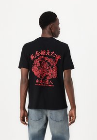 Čierne bavlnené tričko s krátkym rukávom s veľkým červeným grafickým vzorom na chrbte, ktorý obsahuje zložené návrhy a japonský text, s voľným strihom.