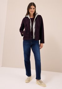 Veste en polaire violet avec un sweat à capuche gris en dessous, associée à un jean bleu et des baskets claires. Texture lisse, poches avant, fermeture à bouton.
