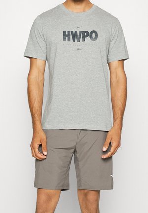 Homme portant un t-shirt gris "HWPO" et un short taupe, debout les bras détendus contre un fond clair.