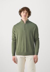 Zeleni zip-up pulover iz mehkega materiala, z dolgimi rokavi, rebrastimi manšetami in ravnim robom. Model stoji ob nevtralnem ozadju.