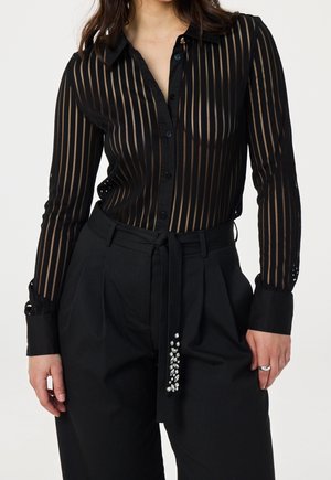 Vrouw met een zwarte doorschijnende blouse met knopen en verticale strepen, gecombineerd met een zwarte broek met hoge taille en een ceintuur met juweelaccent.