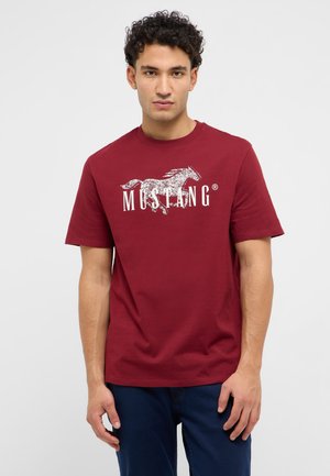 Junger Mann mit lockigem Haar, der ein rotes T-Shirt mit einem weißen laufenden Mustang-Motiv und Text trägt, vor einem schlichten Hintergrund stehend.