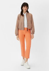 Brązowa i kremowa bomberka w wzór houndstooth, biała koszula zapinana na guziki, pomarańczowe, krótkie dżinsy i białe trampki. Akcenty z elastycznymi mankietami.