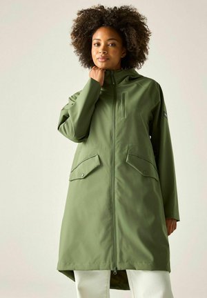 Vrouw met krullend haar die een knielange olijfgroene jas draagt met voorzakken en een ritskraag, staand tegen een effen achtergrond.
