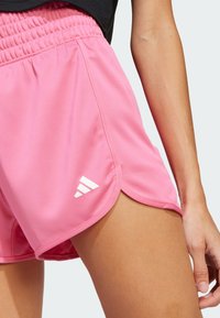 Ružičaste atletske kratke hlače s glatkom teksturom, elastičnim pojasom i bočnim prorezom. Na donjoj lijevoj strani se nalazi bijeli Adidas logo.