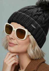 Black knitted beanie with braided texture and a pom-pom on top, paired with large white sunglasses featuring brown tinted lenses.