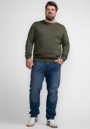 Olive green knit sweater with round neck, paired with blue jeans. Model wears white and black sneakers. Smooth texture, fitted design.
