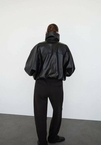 Chaqueta de cuero negra de gran tamaño con cuello alto, combinada con pantalones negros. Textura suave, corte holgado, detalles de costura visibles.