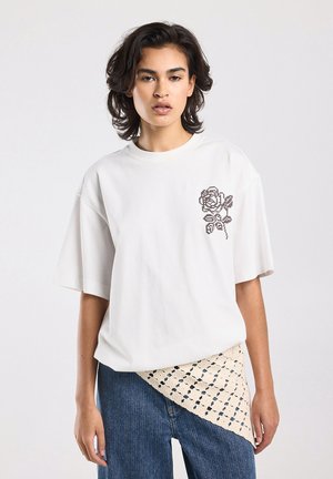 Jong persoon met kort donker haar, gekleed in een wit oversized T-shirt met een gestikte roosontwerp en een spijkerbroek met een crèmekleurige doek met patroon overheen.