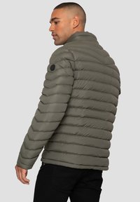 Olivgrüne Pufferjacke mit horizontaler Steppung, Stehkragen und einem runden Logopatch auf der linken Schulter. Strukturiertes Material.