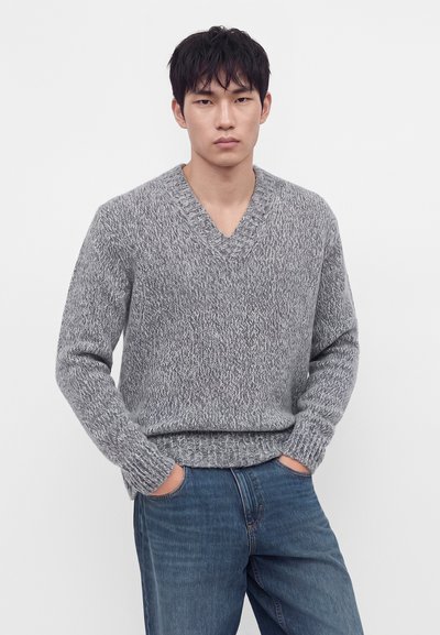 Jeune homme aux cheveux noirs portant un pull gris en maille à col en V et un jean bleu, les mains dans les poches, sur un fond uni.