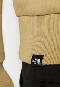 Suéter de color marrón claro con cuello ajustado. La etiqueta negra presenta el logo "The North Face". El tejido texturizado contrasta con los pantalones oscuros.