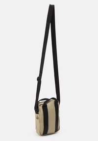 Bézs színű vászon keresztpántos táska fekete pántokkal. Felső fogantyúkkal és oldalsó zsebbel rendelkezik. Téglalap alakú, cipzáras záródással.