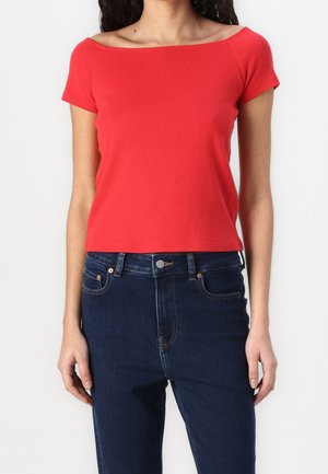 Rotes, kurzärmeliges, schulterfreies Oberteil aus weichem Material, mit tailliertem Schnitt und cropped Länge, kombiniert mit dunkelblauen Jeans.