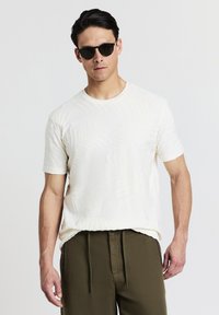 T-shirt bianco a maniche corte con un motivo a foglia testurizzata, abbinato a pantaloni verde oliva e completato da occhiali da sole neri a forma rotonda.