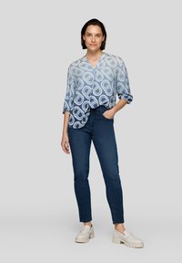 Hellblaue Bluse mit großen weißen geometrischen Mustern, Dreiviertelärmel und lockerer Passform, kombiniert mit dunklen Jeans und hellen Schuhen.