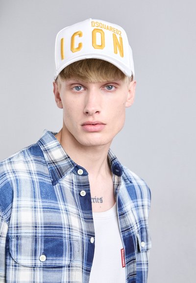 Jaunas vyras su mėlynomis akimis, dėvintis baltą kepurę su geltonu užrašu "ICON" ir mėlyną languotą marškinį virš baltos camisole prieš šviesų foną.