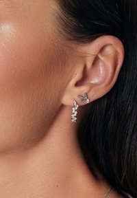 Srebrne kolczyki z liściastym wzorem i zwisającymi kryształami; zawierają wiele przezroczystych kamieni szlachetnych oraz teksturalny akcent liścia.