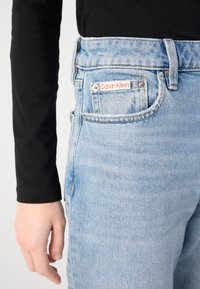 Lys blå denimbukser med høy midje, frontlommer og et Calvin Klein-merke på baklommen. Teksturert stoff med en falmet finish.