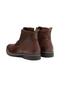 Botas de cuero marrón hasta el tobillo con un acabado texturizado, cordones en la parte delantera y cremallera lateral. Presentan una suela de goma negra y un acento rojo en contraste.
