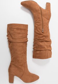 Botas altas hasta la rodilla de ante color camello con diseño holgado, punta puntiaguda y cremallera lateral. Tacón grueso que ofrece estabilidad. Textura lisa en todo el calzado.