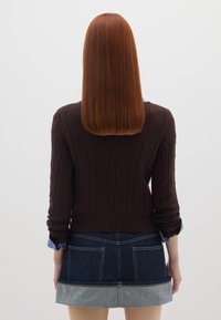 Maglione marrone lavorato a maglia con texture a coste e dettagli a trecce, abbinato a una mini gonna in denim con orlo a contrasto e tasche.