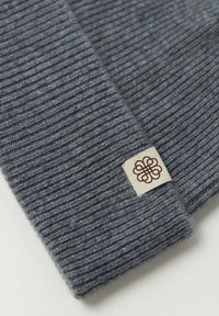 Grå stickad mössa med ribbad textur och en synlig tygetikett med en brun logotyp. Materialet verkar mjukt och varmt.