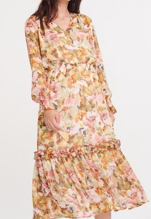 Vrouw draagt een midi-jurk met lange mouwen, bloemenpatroon, elastische taille en ruchesdetail bij de zoom in zachte roze, gele en beige tinten.