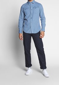 Ljusblå långärmad skjorta med knappar och två bröstfickor, ihop med mörka byxor och vita sneakers. Har en rundad nederkant och krage.