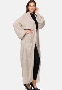 Beige knitted cardigan with a loose fit, long sleeves, and a patch pocket. The texture is chunky, providing a cozy appearance.