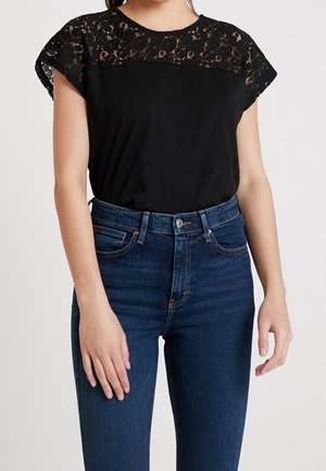 Kvinde iført en sort blonder kortærmet top kombineret med højttaljede blå jeans, stående mod en ensfarvet lys baggrund.