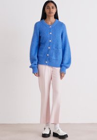 Blauer gestrickter Cardigan mit Rundhalsausschnitt, übergroßen Ärmeln und großen Knöpfen; kombiniert mit hellrosa Hosen und zweifarbigen Schuhen.