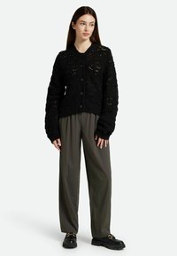 Cardigan noir en maille avec un motif intricate, fermeture à boutons et manches surdimensionnées. Associé à un pantalon à rayures taille haute et des chaussures noires massives.