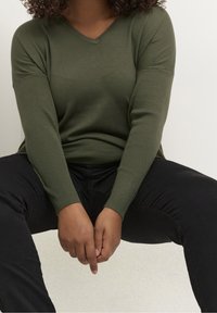 Grøn v-hals sweater med teksturerede ribstrikkede ærmer, tætsiddende pasform og en blød finish, parret med sorte bukser. Hænderne hviler på knæene.