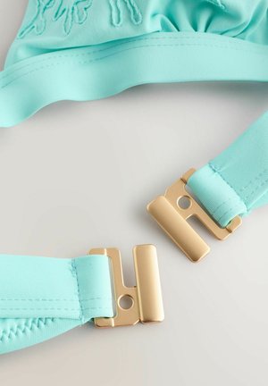 Šviesiai mėlyna bikini viršutinė dalis su surinkta medžiaga ir auksiniais metaliniais užsegimais. Lygus paviršius su siuvimo detalėmis ant dirželių ir medžiagos kraštų.