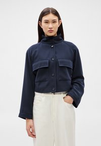 Femeie care poartă o jachetă scurtă bleumarin cu nasturi, buzunare mari și guler înalt, asortată cu pantaloni albi cu talie înaltă.