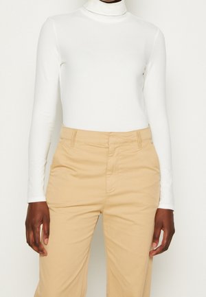 Personne portant un haut à col roulé blanc à manches longues et un pantalon beige taille haute, montrée du cou aux hanches sur un fond uni.