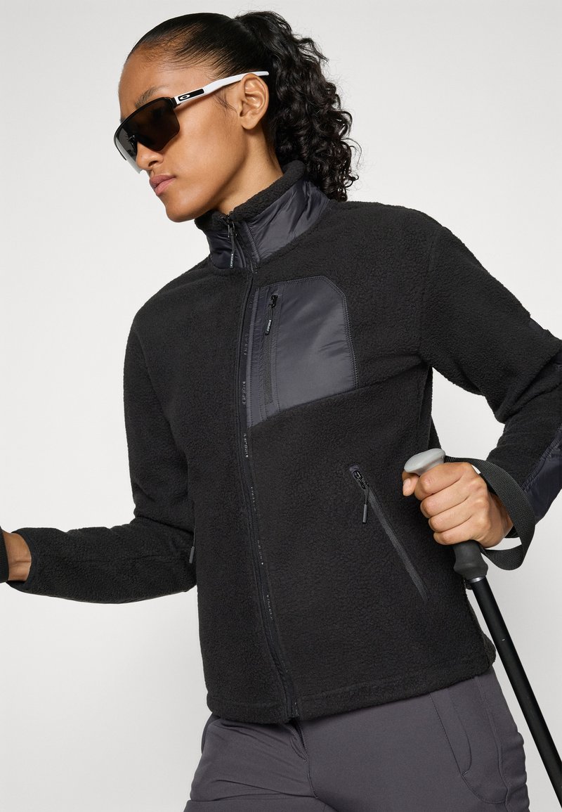 Eine Frau trägt eine schwarze Fleecejacke und dunkle Sonnenbrillen, hält Skistöcke und bereitet sich auf eine Outdoor-Aktivität vor, vor einem weißen Hintergrund.