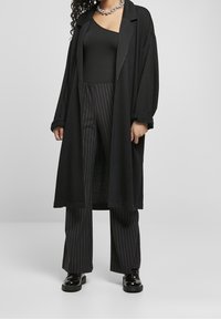 Cardigan lungo nero con una vestibilità morbida, apertura frontale e polsini arrotolati, abbinato a pantaloni neri a righe e scarpe con piattaforma nere lucide.
