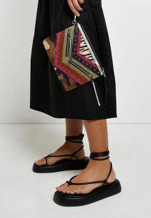 Henkilö, jolla on mustat hihnalliset korokepohjasandaalit ja musta mekko, pitäen moniväristä geometrisella kuviolla varustettua clutch-laukkua, jossa on vetoketjut.