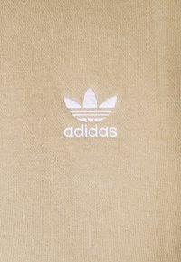 Bílý vyšívaný trifold logo Adidas na béžové pletené tkanině, umístěný na středu a jasně viditelný.